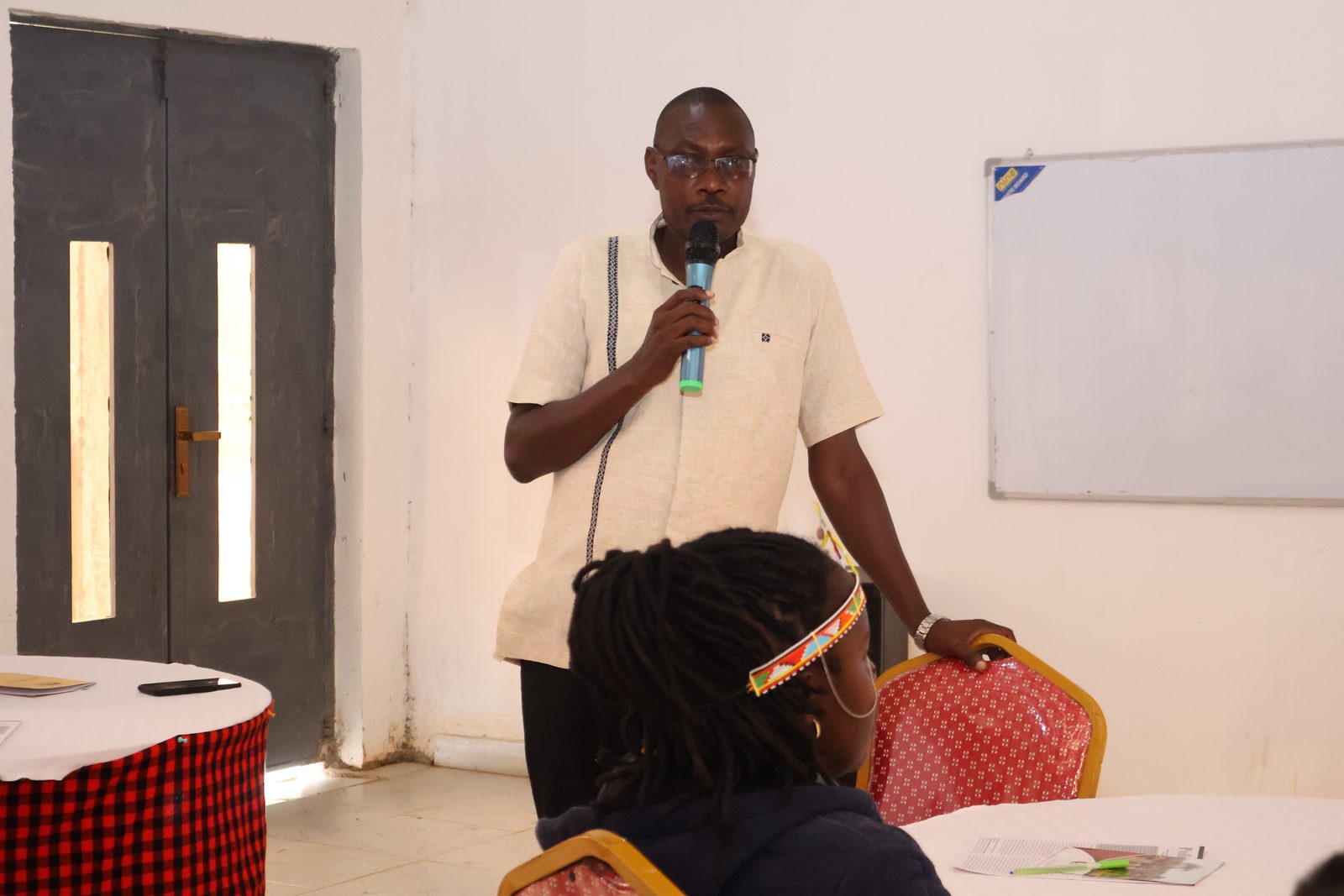 A man stands and speaks into a microphone during a workshop, a seated participant in a beaded headband listening in front of him.