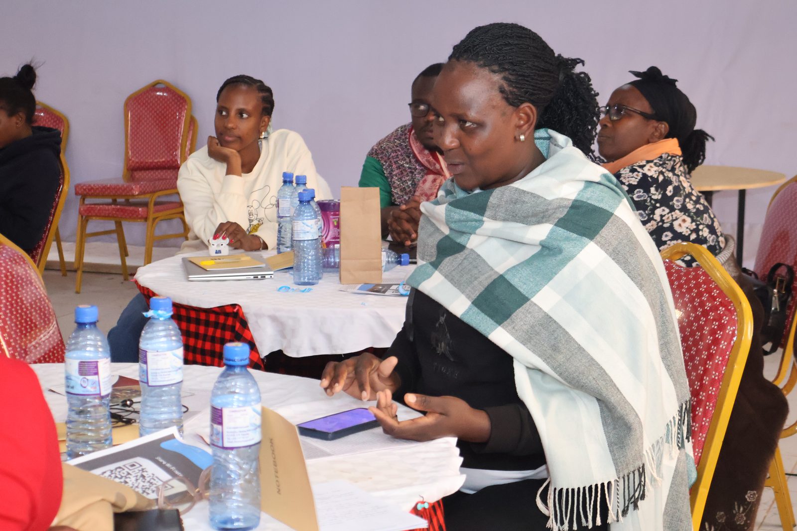 A woman wrapped in a checked shawl speaks and gestures while colleagues listen at a discussion table.