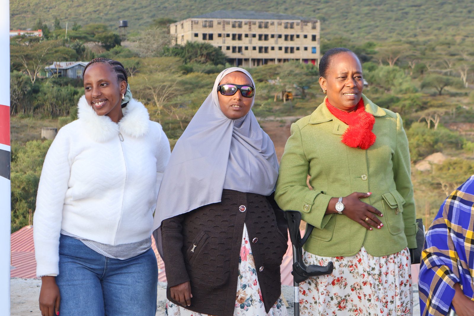 Three women stand together outdoors at sunset, one steadying herself on a forearm crutch, smiling toward the group.