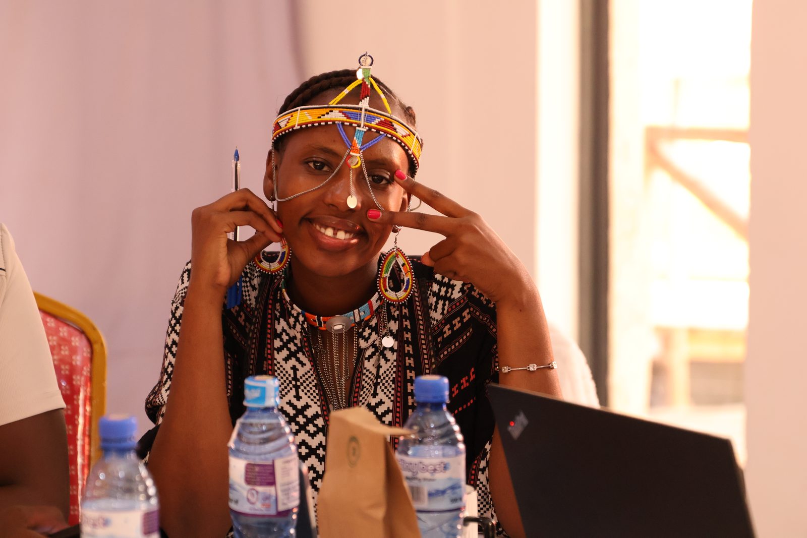 A participant in a beaded Maasai headdress and patterned dress smiles warmly at a workshop table beside a laptop.